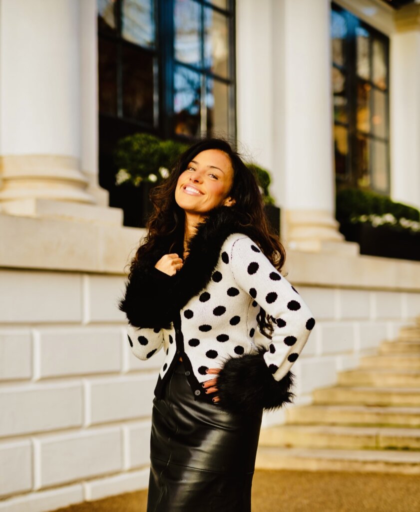 Laura White posing in front of a building, outside, wearing a white with black polka-dot jumper-jacket with faux-fur collar. She's beaming with happiness and wearing a black skirt underneath her jacket.