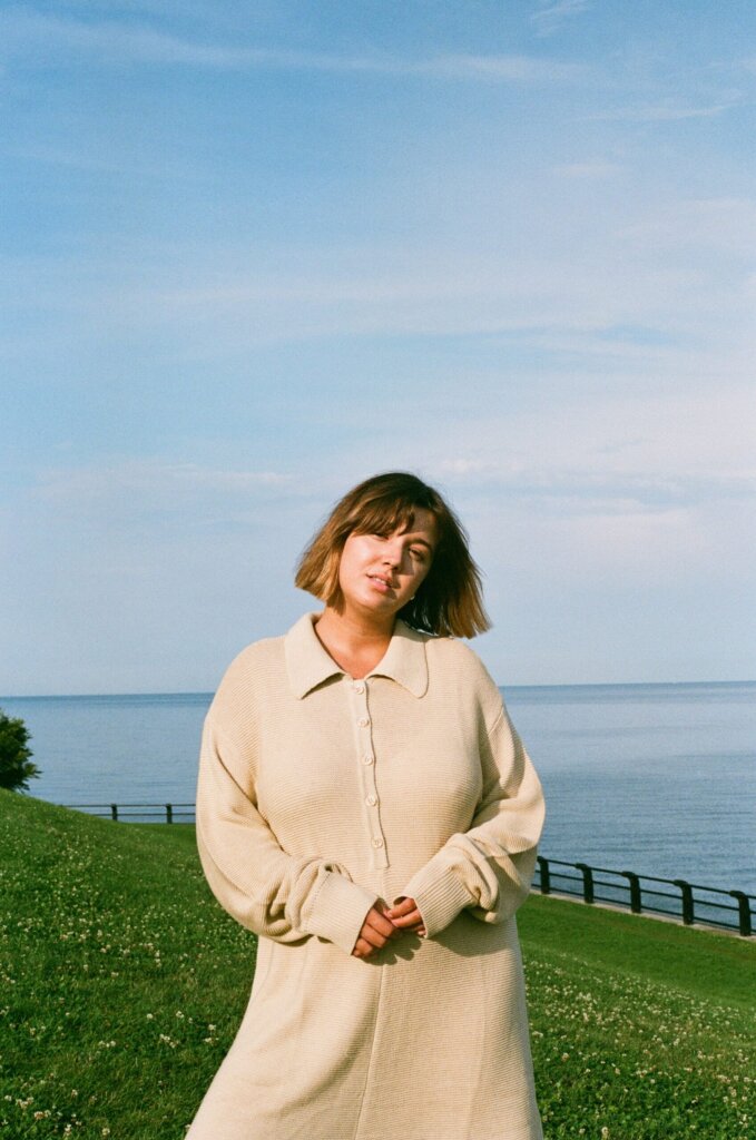 Alle The Dreamer in a promotional photo for "Run Home To You", which sees her standing in a beige outfit on a green hill with the ocean behind her.