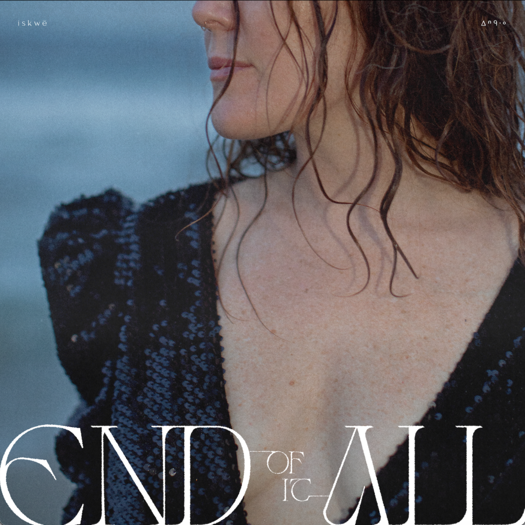 Official single cover artwork for "End Of It All" which is sees the camera focus on iskwē's cleavage as she turns her head to the side while wearing a black dress on a cold beach.