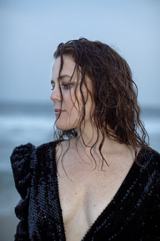 Promotional photo for "End Of It All" which sees a mid-shot of iskwē wearing a black dress with a v-neckline that reaches her midriff, showing off her cleavage. She has her head turned to the side and her auburn hair is slightly wet. She looks like she's on a cold beach.