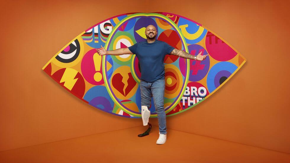 Big Brother 2023 housemate Dylan standing in front of the new eye logo with a bright orange background.