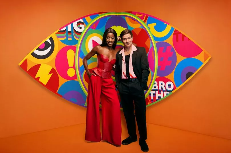 The hosts, AJ Odudu and Will Best, standing in front of the new Big Brother logo with a bright orange background. AJ is wearing a gorgeous red dress and Will is wearing a black suit with a pink shirt that is opened to show a black vest.