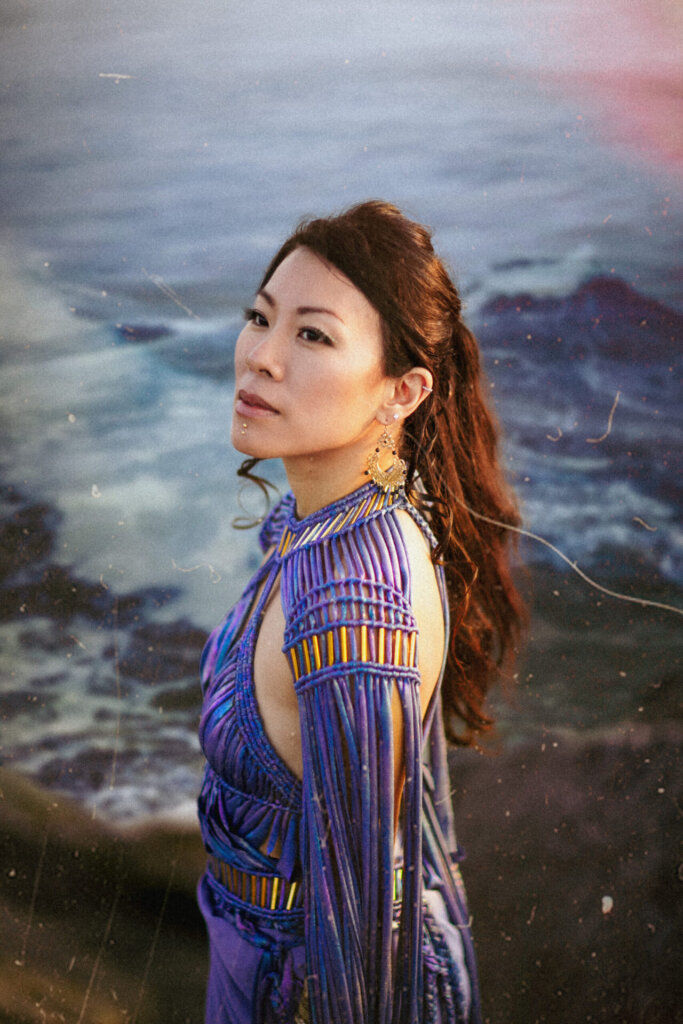 Promotional photo for "Cobalt Sea", which sees Eunice Keitan standing to the left with her head turned to the camera, wearing a blue dress that matches the sea behind her.