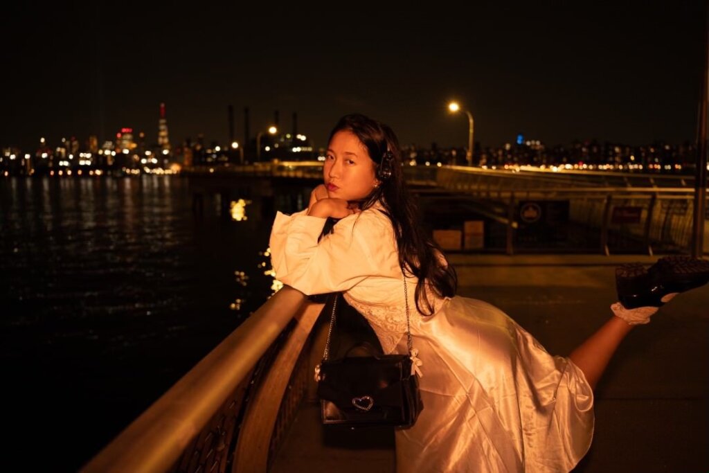 Promotional image for "VIDEO CALL: PM mini movie" which sees ÊMIA posing with the New York skyline behind her, wearing a cream silk skirt, a white top, accessorised with a black bag and black shoes. She's leaning against a rail, overlooking the water, in the night time, with one leg pointed up behind her - bent at the knee.