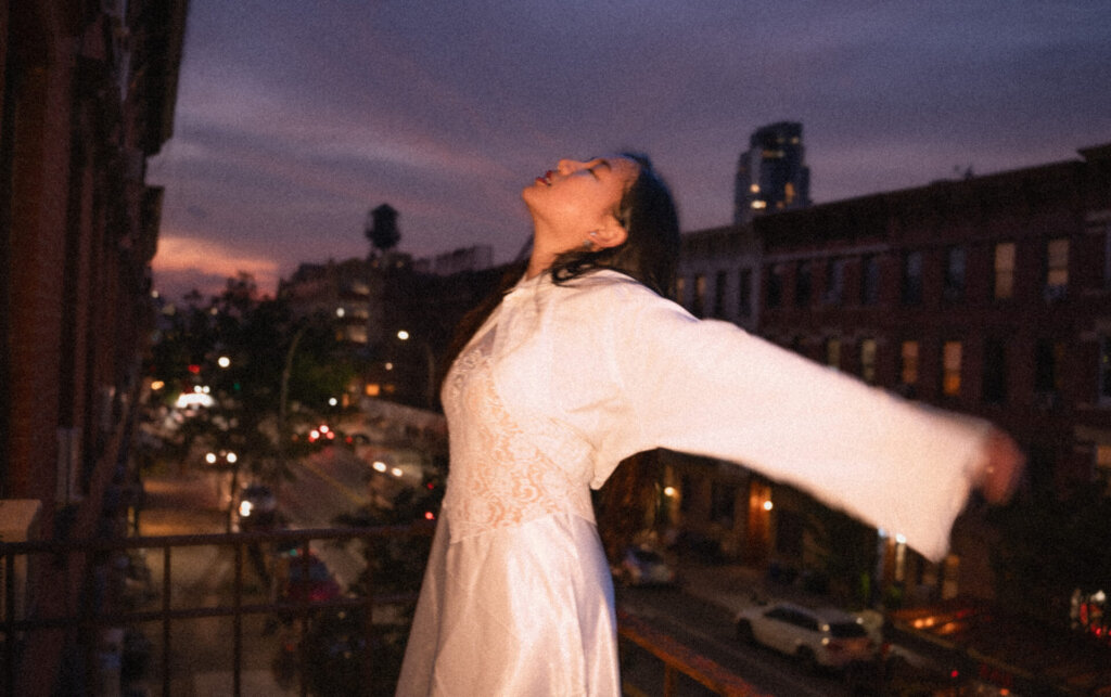 Promotional image for "VIDEO CALL: PM" which sees ÊMIA posing with her arms open wide with the New York skyline behind her as the sun sets or rises.