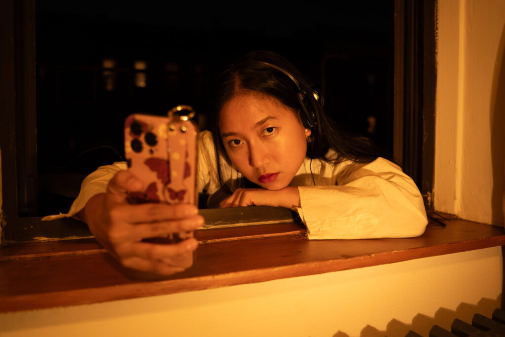 Promotional image for "VIDEO CALL: PM" which sees ÊMIA posing in a window frame, with her arm perched on the windowsill, looking right at the camera, with her phone out in front of her, taking a selfie.