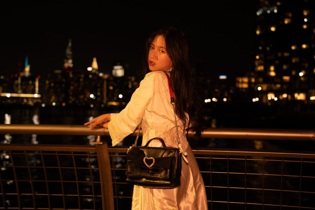 Promotional image for "VIDEO CALL: PM" which sees ÊMIA posing against a rail, with the New York skyline behind her, looking at the camera over her shoulder. She's wearing a white top and a silk skirt with a black bag and it's night time.