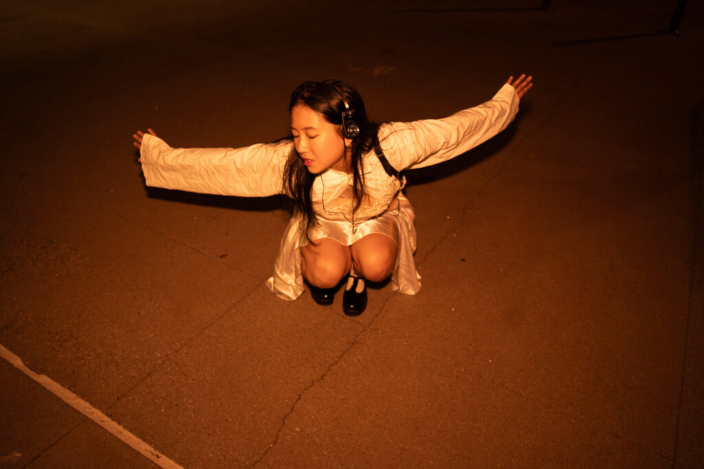 Promotional image for "VIDEO CALL: PM" which sees ÊMIA posing by crouching down in a white top and a cream silk dress as she flings her arms open for the photo.