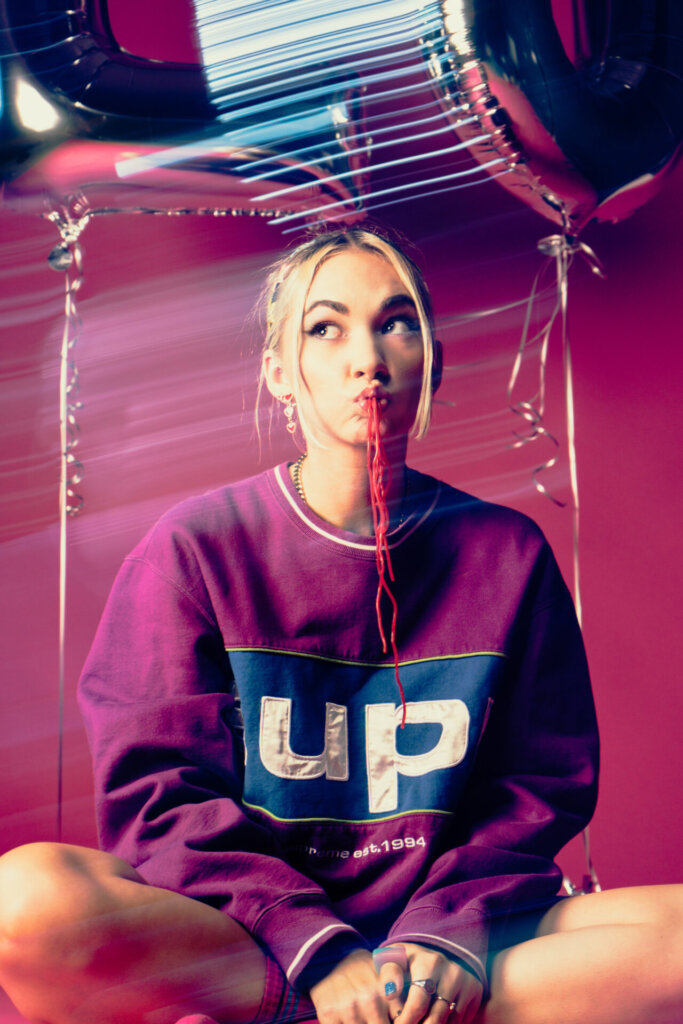 Promotional photo for "Monster" which sees Lo Lauren wearing a pink sweatshirt, sitting crossed legged on the floor, eating red sweet laces.