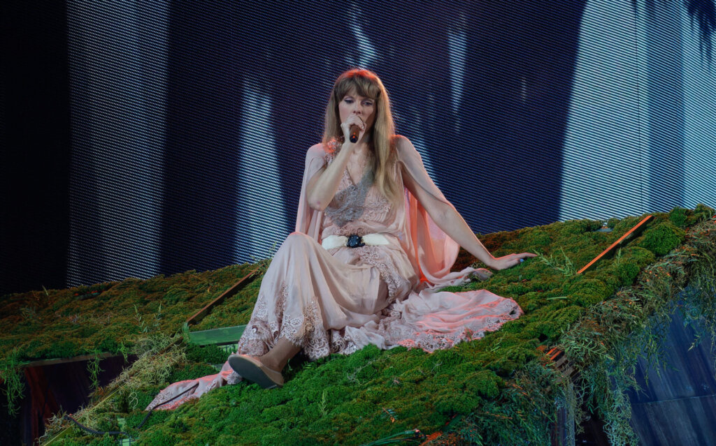 Taylor Swift sitting on a grassy hill performing on stage wearing a pale pink dress.