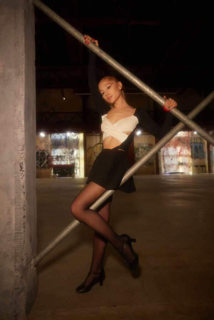 Ariana Grande posing for a promotional image for "yes, and?" wearing a black skirt and a white cross top with her blonde hair in a ponytail. She's posing against two grey poles.