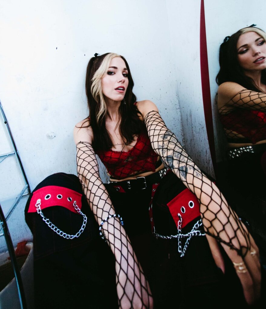 Promotional photo for "IDWTAY" which sees Dela Kay posing against a mirror, wearing black and red trousers with a red bra top and she has black hair with two front blond streaks.
