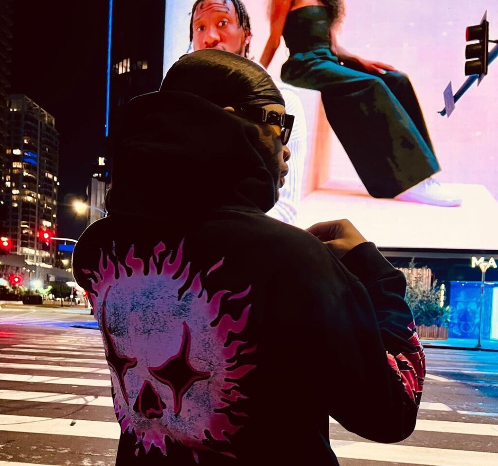 Promotional image which sees Just Listen crossing a road with his back to the camera showing off his hoddie with a skull print on the back. There's a huge LED screen in front of him.