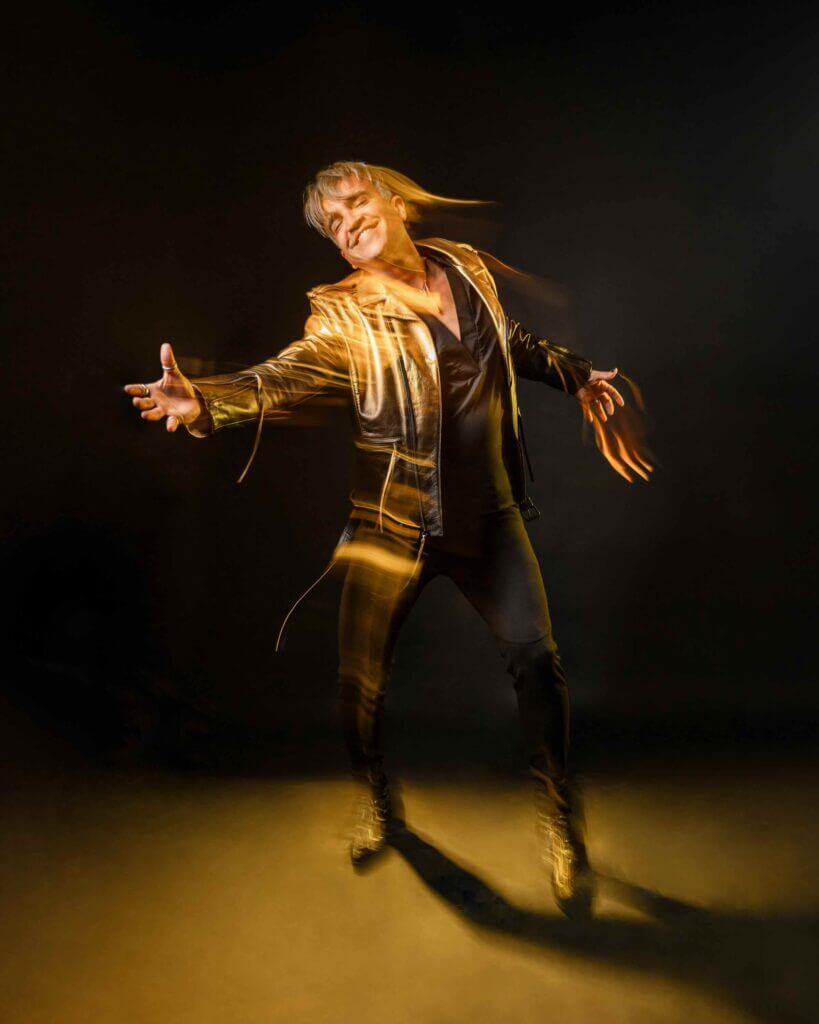 Promotional photo for "Kaleidoscope" in collaboration with Velvet Code which sees a gold-yellow-filtered shot of Roger Kuhn, standing and reaching out in a gold-encrusted suit and matching trousers that is photographically distorted.