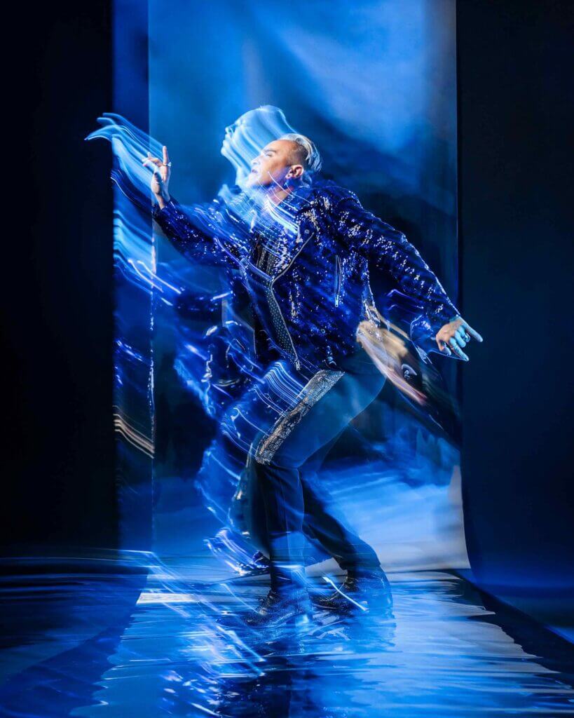 Promotional photo for "Kaleidoscope" in collaboration with Velvet Code which sees a sapphire-blue-filtered shot of Roger Kuhn, standing and reaching upwards in a blue-sequined jacket and matching trousers that is photographically distorted.