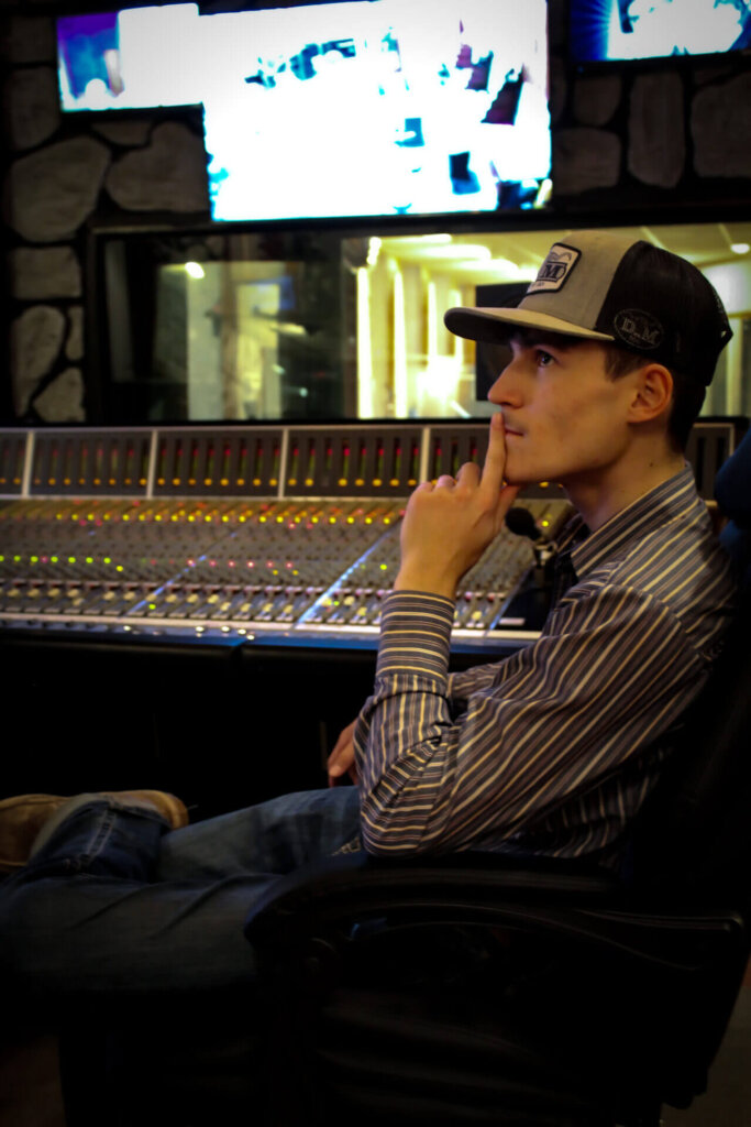 Promotional photo for "Whiskey Talks" which sees Caleb Montgomery in a brown-and-white striped shirt, jeans and a baseball cap, sitting in a producer's chair in a studio.