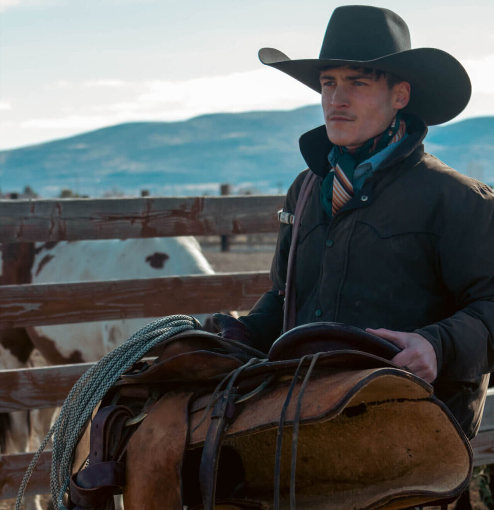 Promotional image for "Whiskey Talks" which sees Caleb Montgomery holding a saddle as he walks past a cow, wearing a jacket and a cowboy hat.