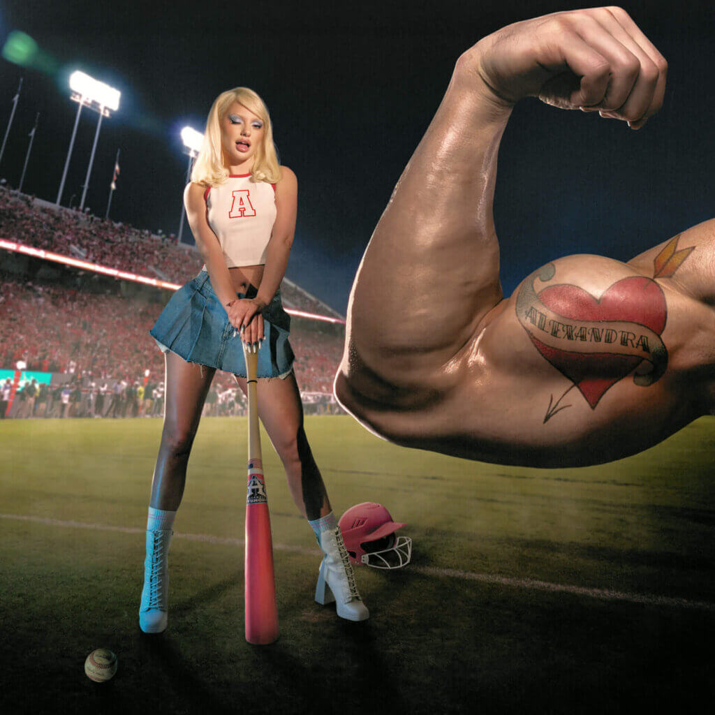 Official single cover artwork for "Băieții" which sees Alexandra Stan in a baseball outfit with a baseball bat on a baseball field with a men's muscular arm tensed up in the foreground which is adorned with a heart tattoo with the name "Alexandra" wrapped around it, on his bicep.