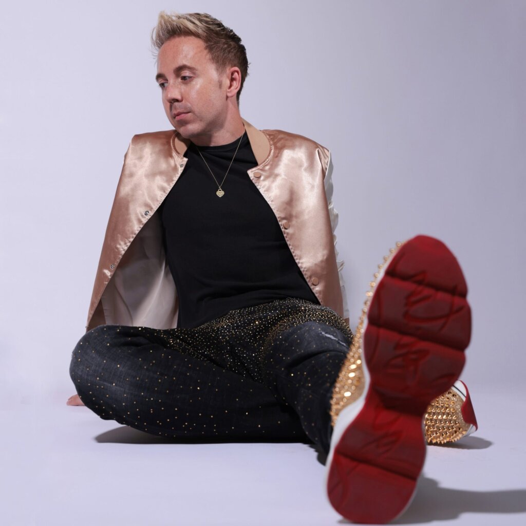 Press photo for John Galea which sees him sitting on the floor posing for a photo, wearing a shiny gold jacket, a black t-shirt, glittery black jeans, and a trainer with a red sole.