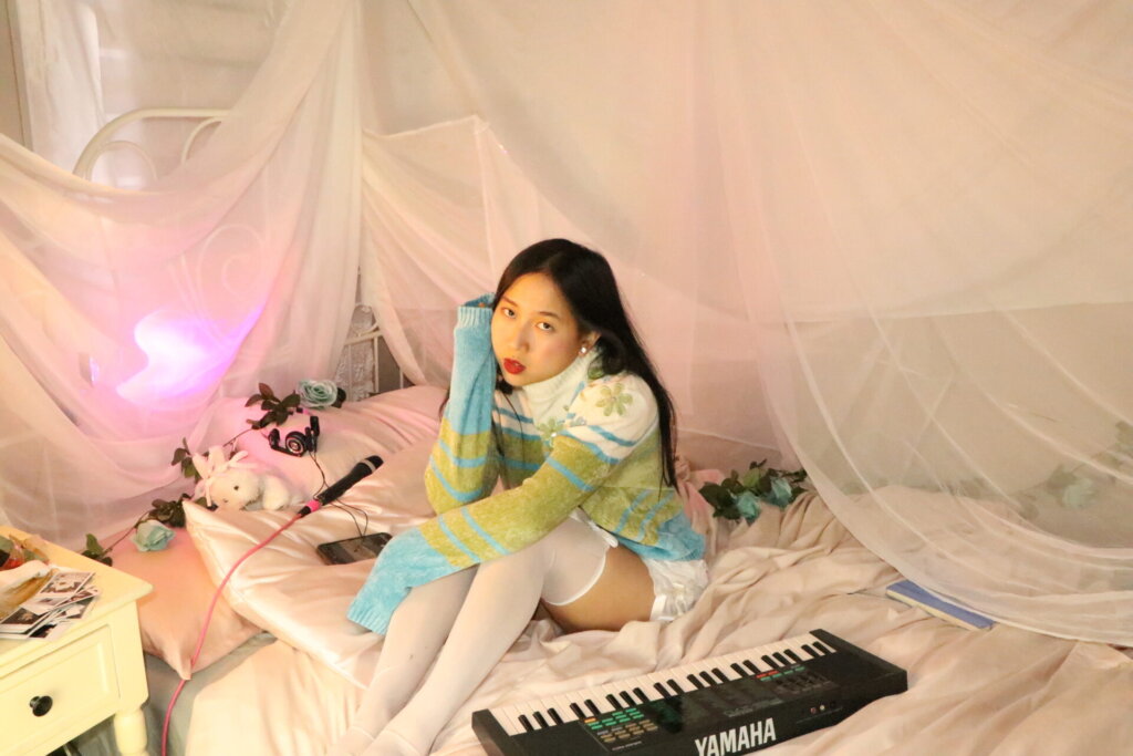 Promotional photo for "Super Fun Party Girl" which sees ÊMIA sitting on a bed with a keyboard out in front of her she hugs her knees, looking at the camera with her dark hair behind her, wearing a white-to-green-to-blue jumper and white stockings. There's a pink hue net over the bed and some small teddies on her pillow.