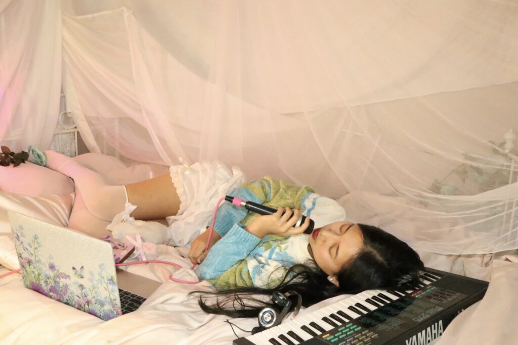 Promotional photo for "Super Fun Party Girl" which sees ÊMIA lying on a bed with a keyboard out in front of her as she rests her head on the keyboard, singing into a microphone with her eyes closed. Her dark hair is on the keyboard and she is wearing a white-to-green-to-blue jumper and white stockings. There's a pink hue net over the bed and some small teddies on her pillow. She has a laptop out in front of her.