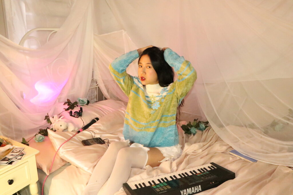 Promotional photo for "Super Fun Party Girl" which sees ÊMIA sitting on a bed with a keyboard out in front of her as she puts her hands on the top of her head and stretch, she's looking at the camera with her dark hair falling behind her. She is wearing a white-to-green-to-blue jumper and white stockings. There's a pink hue net over the bed and some small teddies on her pillow.