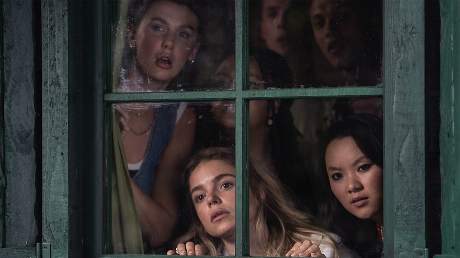 Still from Wreck TV series that sees a group of young adults looking out of a window as if they are hiding from someone.