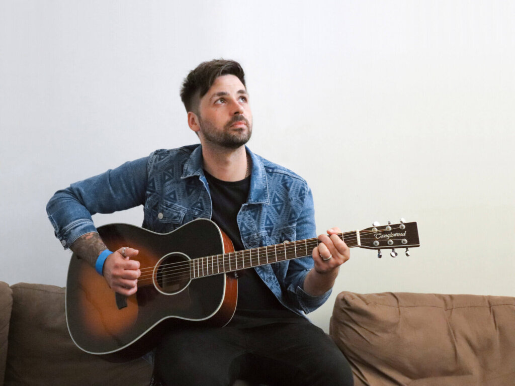 Promotional photo for "Easy (Like Sunday Morning)" which sees Ben Haenow sitting on a couch with a guitar across his lap, earing a denim jacket and black skinny jeans.