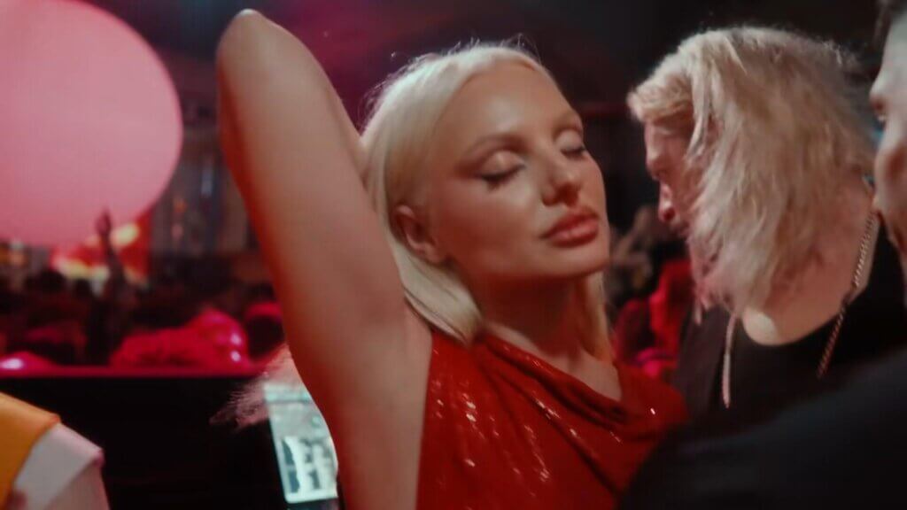 Still from the "Băieții" music video which sees Alexandra Stan feeling herself at the DJ booth, dancing to music, with her eyes closed and one arm behind her head.