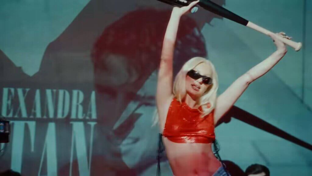 Still from the "Băieții" music video which sees Alexandra Stan performing on stage wearing a red top and sunglasses, holding a baseball bat above her, with a projection of boys behind her.