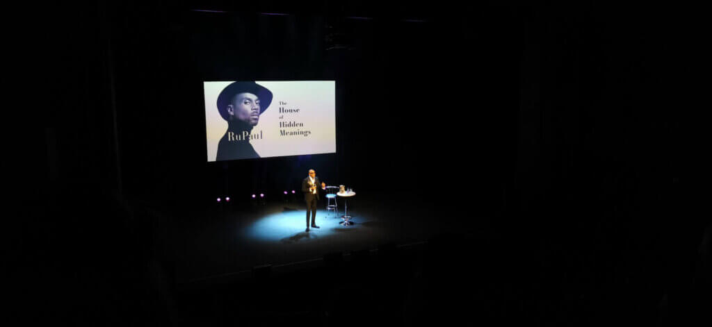 RuPaul wearing a black suit, standing on stage with a table and chair to his left and a projection of the cover image of "The House of Hidden Meanings" book behind him.