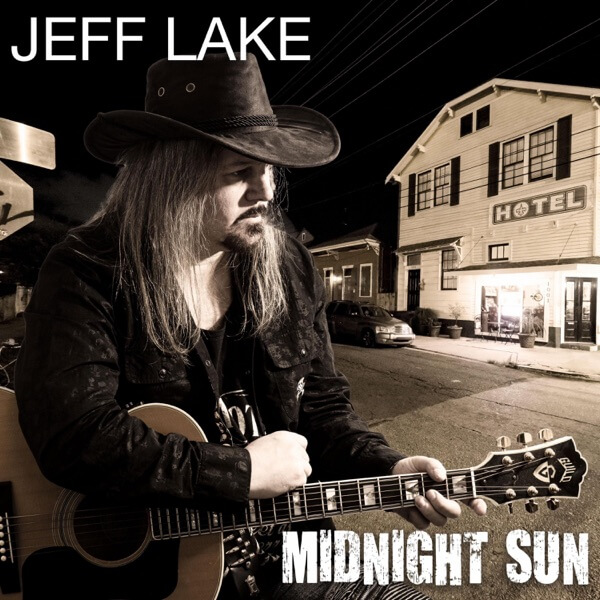 Album cover artwork for "Midnight Sun" which sees Jeff Lake posing with his guitar outside and across the street from a hotel, wearing a dark jacket and a dark cowboy hat. The image has a sepia black and white filter to it.