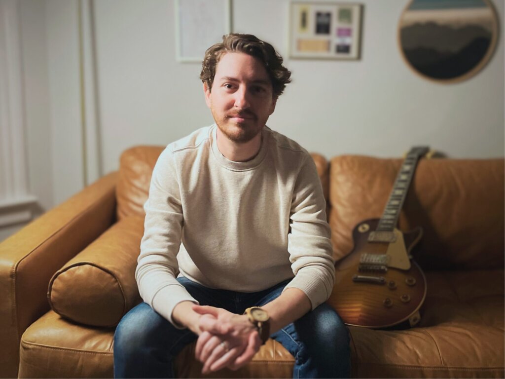 Promotional image for "When The Lights Go Down" which sees Kevin Harrison sitting on a brown couch with a guitar behind him, leaning forward with his hands touching and his elbows on his knees. He is wearing a cream jumper and blue jeans.