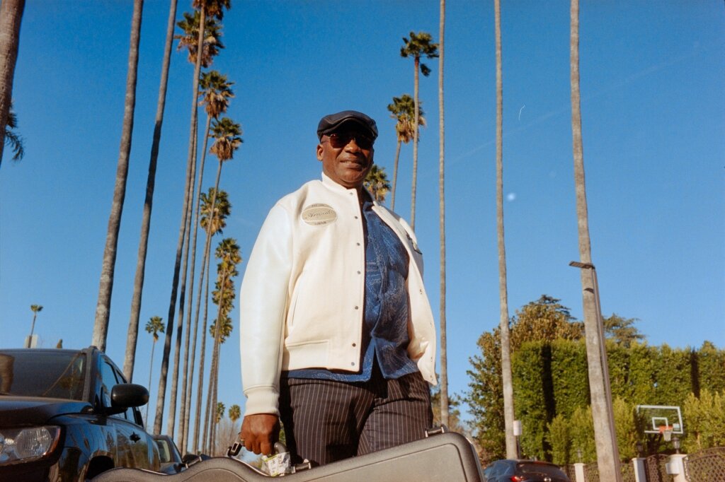 Marius Billgobenson standing on what looks like a typical Beverly Hills road, holding a guitar case, wearing a white jacket and a blue shirt as well as some shades.