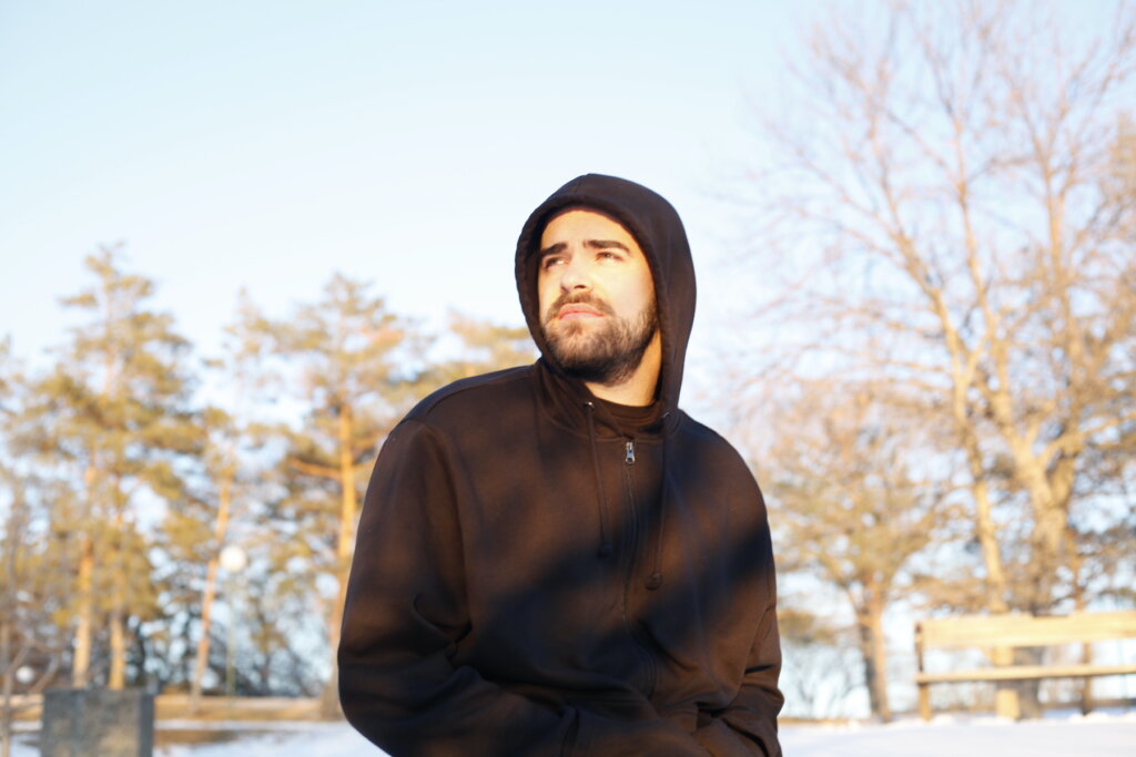 Ben Sefton outside wearing a black hoodie with the hood over his head, looking to the left with his hands in the hoodie's pockets, with trees in the distance behind him.