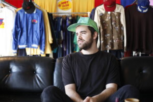 Ben Sefton sitting on a black couch with clothing hung up in what looks like a clothing store, behind him. He's wearing black with a bright green snapback cap, looking off to the left with a thoughtful expression.