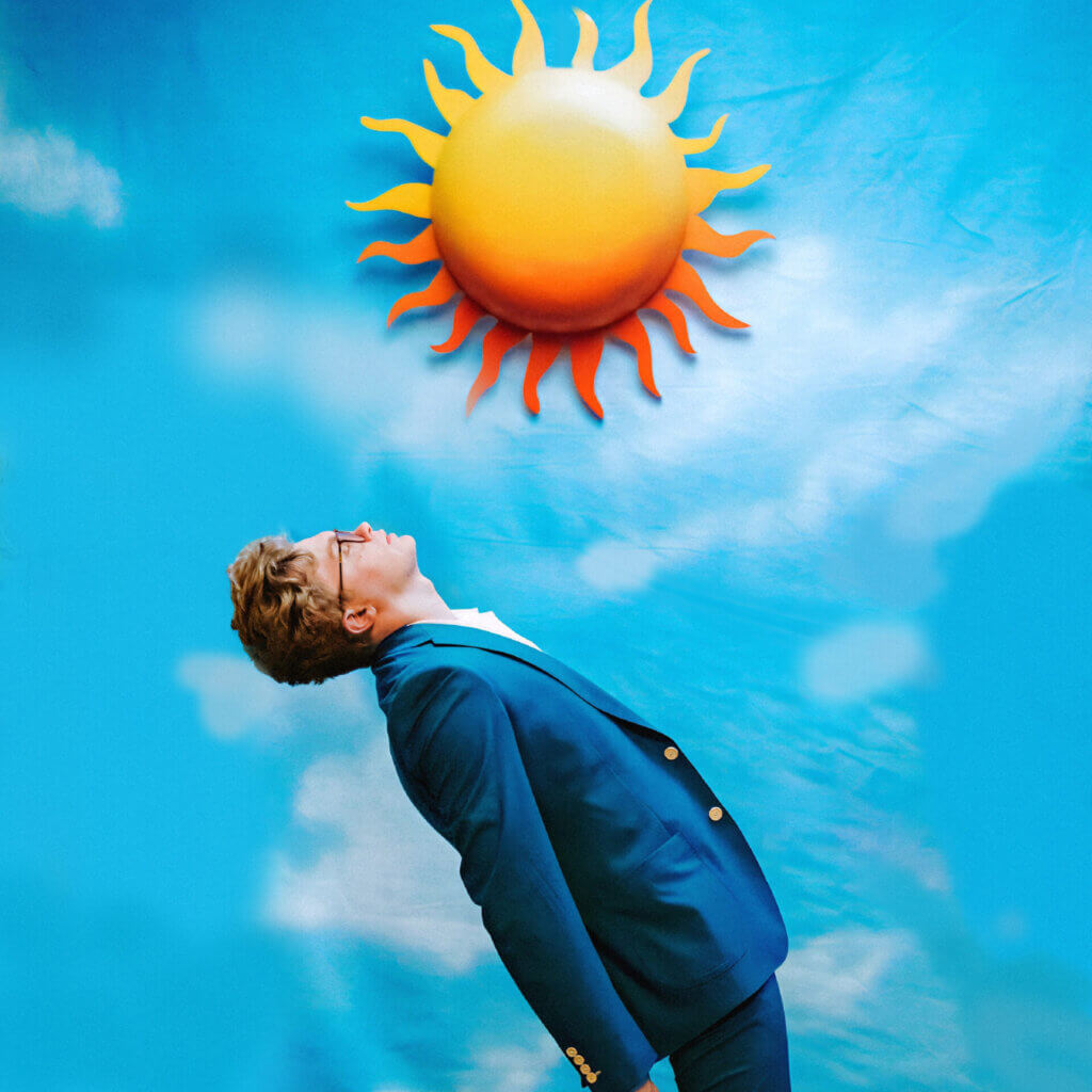 Official single artwork for "Forbidden Feeling" which sees CG5 turned to the right, wearing a blue suit, bending backwards, looking up at a sun emoji that has been enlarged, with a background of clouds.