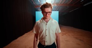 Screenshot from the official music video for "Forbidden Feelings" which sees CG5 standing in a studio wearing an off-white shirt and glasses, with a big blue box behind him.