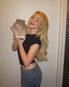 Sabrina Carpenter posing with the golden UK Number 1 chart award trophy for "Espresso" while wearing a black top and some light blue jeans, against a white wall with her eyes closed and her blonde hair falling down her back.
