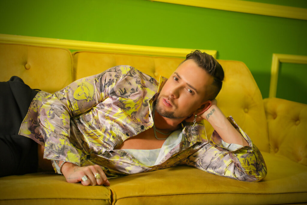 Promotional photo for "I AM" which sees David Hernandez lying down on a mustard-coloured couch wearing a beige and yellow shirt that is unbuttoned halfway revealing his chest. He's also wearing black jeans. He is stretched out on his side with his elbow on the couch with his hand keeping his head up.