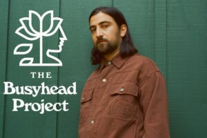 Noah Kahan standing in a brown jacket against a green wall with the logo of The Busyhead Project on the left.