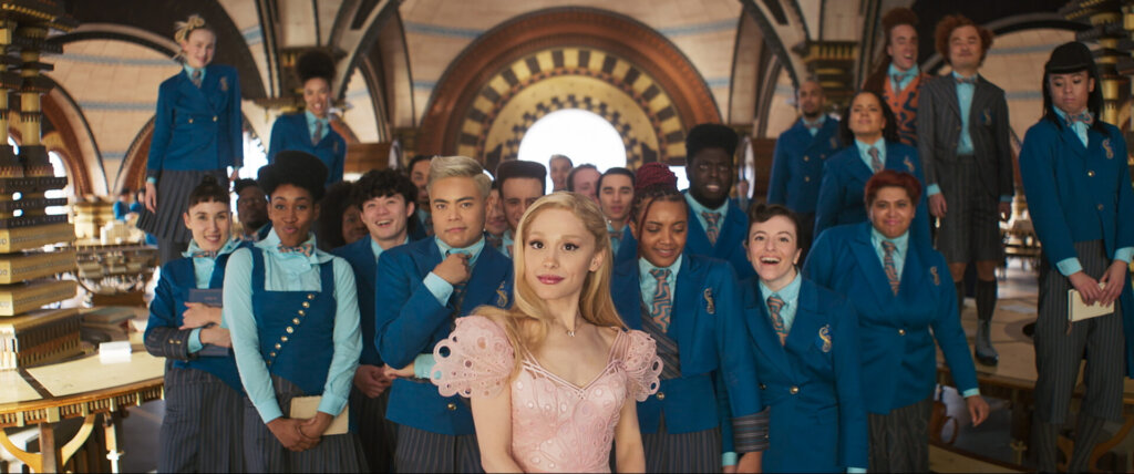 Glinda in front of a popular crowd who are all wearing blue while she wears pink, while they are in a school setting.