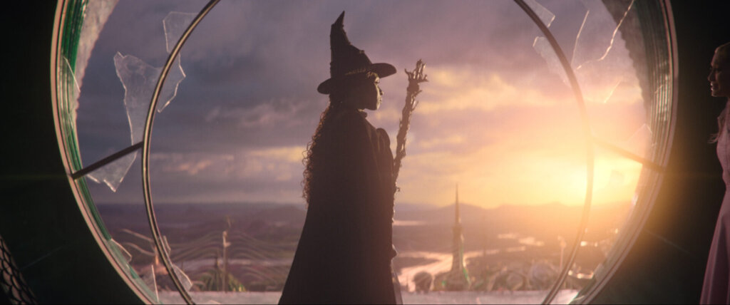 Elphaba as the Wicked Witch of the West, wearing her black clothes and the pointy hat, holding a broomstick as she flies out a circle window.