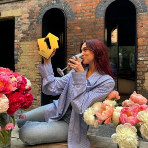 Dua Lipa posing with the gold Number 1 award, holding it in the air, which she got for her "Radical Optimism" album. She's outside with some flowers in front of her, sipping from a glass of wine, wearing a blue shirt and blue jeans.