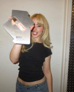 Sabrina Carpenter posing with the golden UK Number 1 chart award trophy for "Espresso" while wearing a black top and some light blue jeans, against a white wall, pulling a grin as she holds the trophy out to the camera.