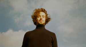 Promotional photo for the live videos of "nothing else" and "flowers" which sees SACHA wearing a brown turtleneck jumper with a cloudy sky behind him.
