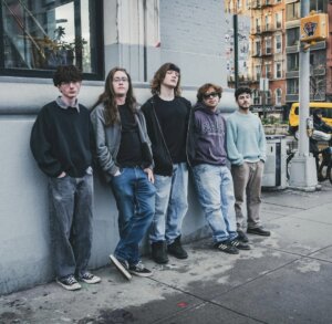 Promotional photo which sees all five band members of SAMSARA. posing against a street wall with a high window, all wearing jeans and hoodies and trainers.