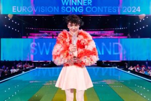 Nemo posing on stage with the glass trophy wearing a baby-pink skirt and a fluffy pink open-chested jacket after winning the Eurovision Song Contest 2024 for Switzerland with the song "The Code".