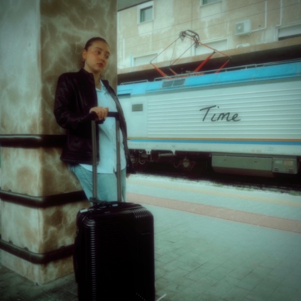 Official single cover artwork for "Time" which sees Maya Ixta leaning against a stone pillar at what looks like a train station, with a black suitcase, wearing light blue jeans, a white top and a black jacket, with her hair tied up. while the word "Time" is displayed on the side of a train. The image has an old-school filter to it.