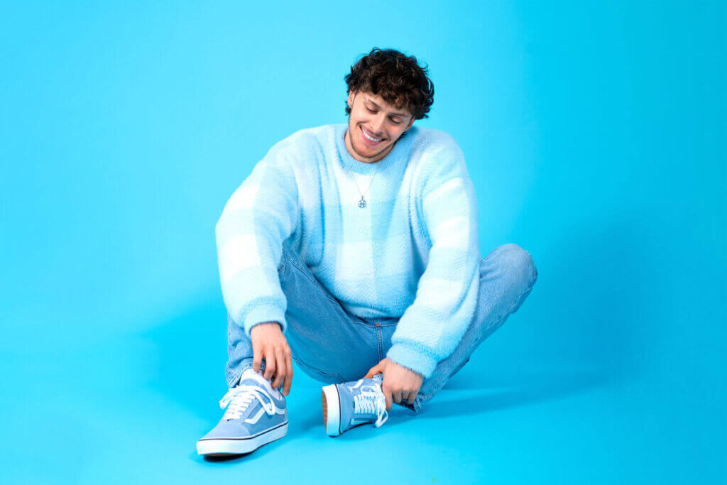 Promotional photo for "Delusional" featuring Mimi Sky, which sees Scott The Pisces sitting on the floor at a photo shoot studio, wearing a baby-blue jumper with a pair of blue jeans and some light blue vans trainers. His background is a bright blue that doesn't match any of the colours he is wearing.